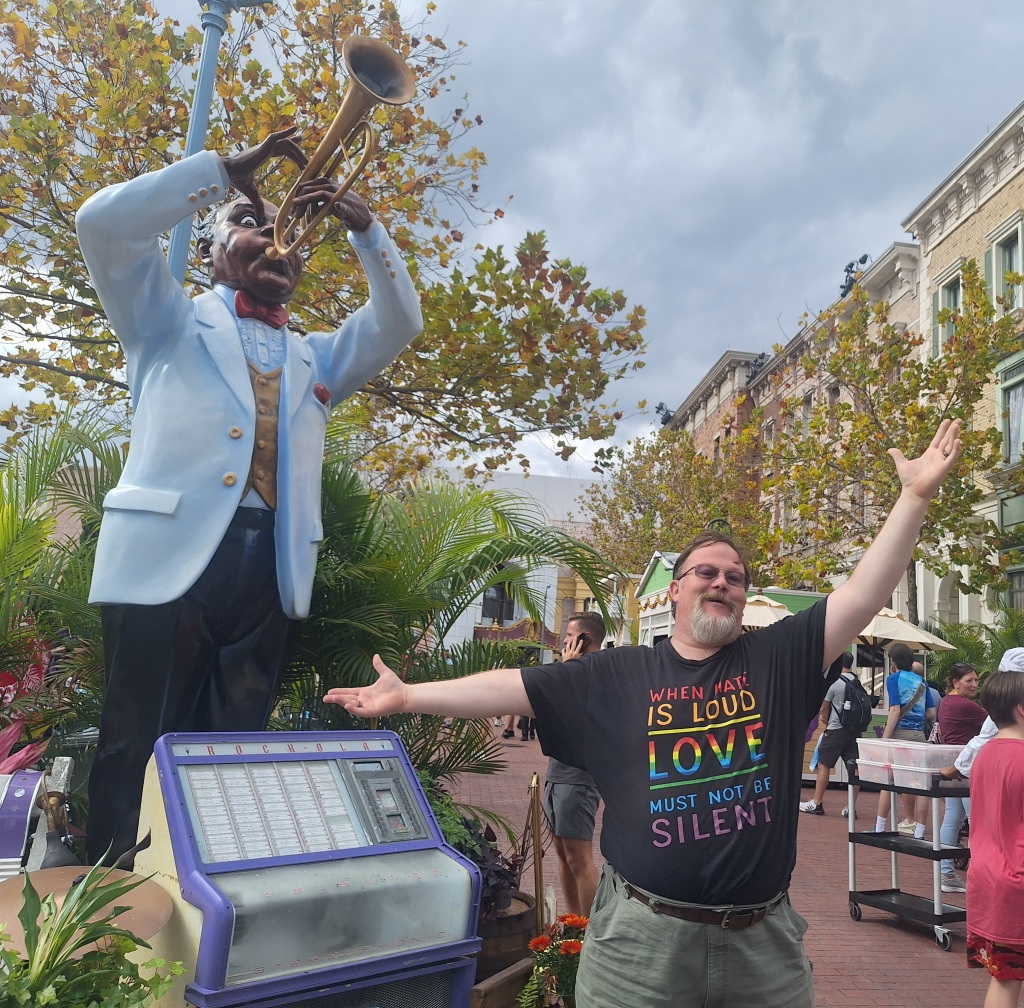 A street scene from within a theme park A tree lined street with old fashioned brick buildings in the background.  Blue sky and clouds above.  In the foreground stands Geoffrey an auburn haired human with dark glasses and a greying beard.  He is striking a dramatic pose with arms outstretched and wears a Tee shirt with the words "when Hate is Loud Love Must not be silent" in rainbow lettering against a black background.  He stands next to a much larger statue/disply of Louis Armstrong playing the trumpet.